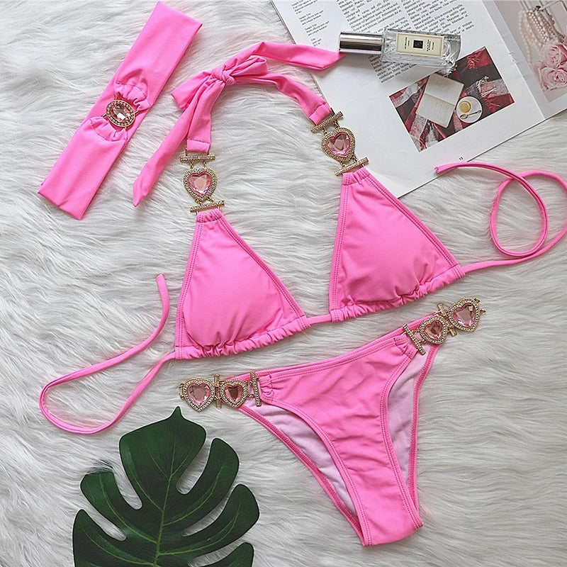 Maillot de Bain Femme Rose Bijoux Cœur, présenté sur un tissu blanc à poil avec un magazine mode, un bandeau assorti au maillot et une feuille de plante verte.